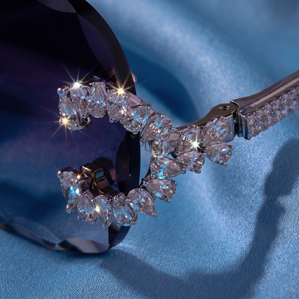 Luxury rimless sunglasses with dark tinted lenses and iced out moissanite stone frames resting on a smooth blue silk background.
