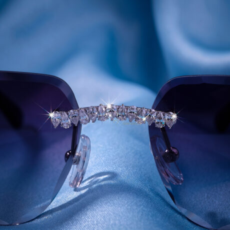 Luxury rimless sunglasses with dark tinted lenses and iced out moissanite stone frames resting on a smooth blue silk background.